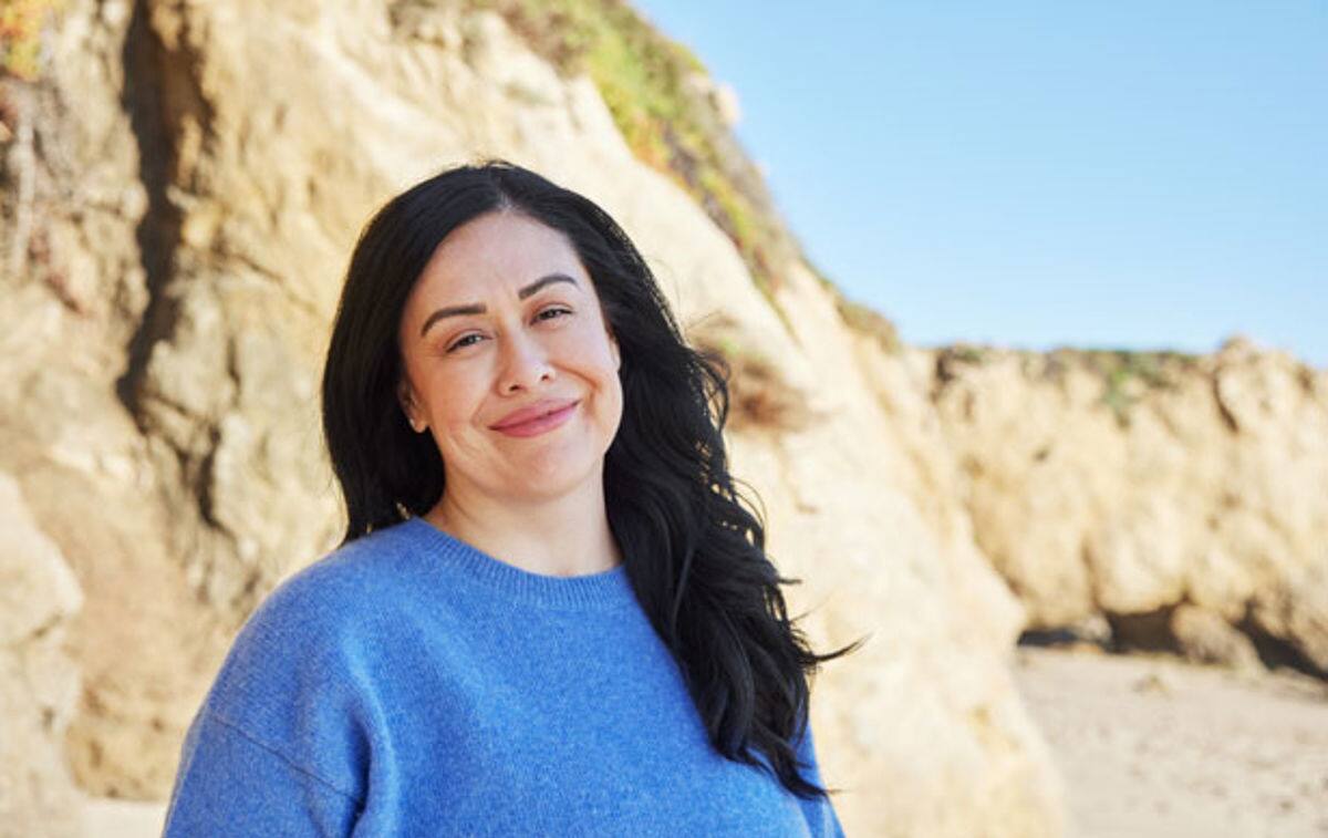 Frau mit schwarzen Haaren und blauen Pulli lächelt vor Steinstrand